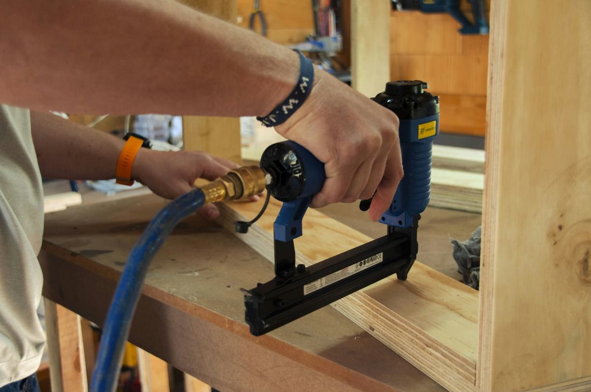 Hands operating a pneumatic brad nailer on a piece of wood.