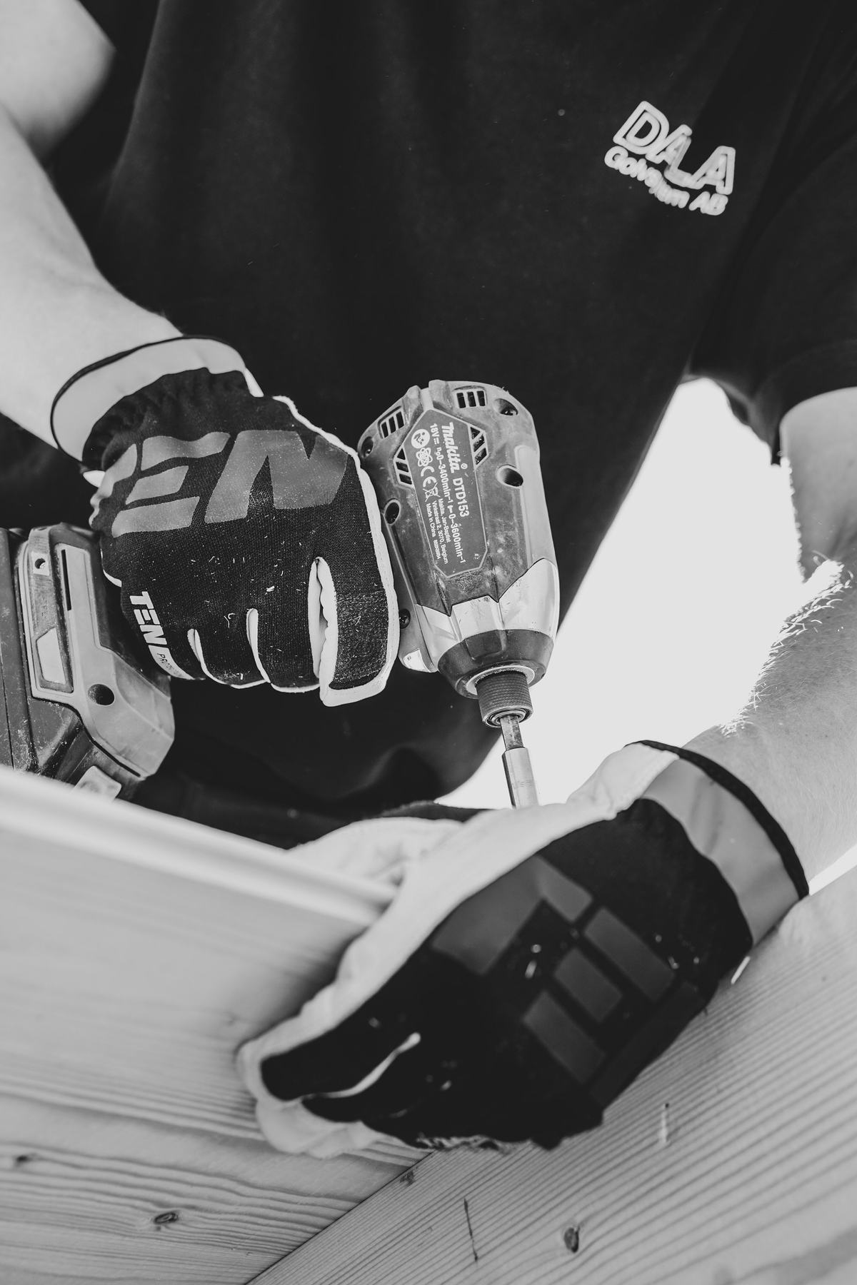 Black-and-white photo of gloved hands driving a fastener into a white panel with an impact driver.