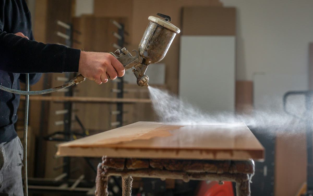 Hand holding an HVLP spray gun applying finish to a flat panel resting on a stand.