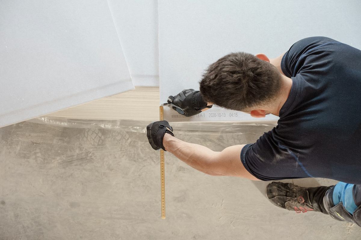 Worker in gloves, seen from above, measuring a bare floor with a folding rule.