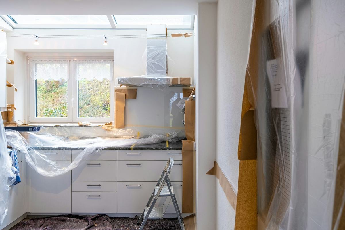 Kitchen under renovation with cabinets and counters wrapped in plastic sheeting and a stepladder on the floor.