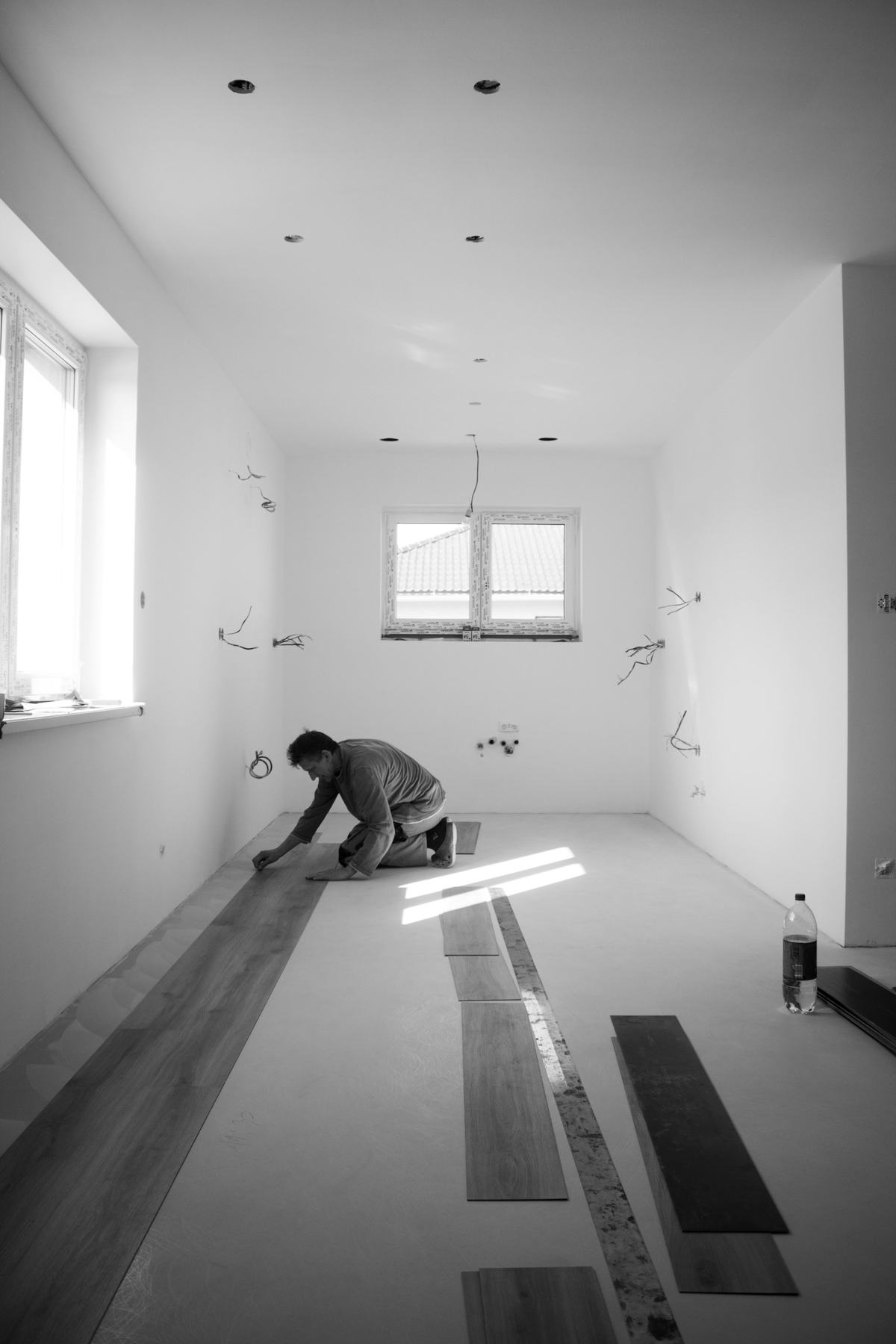 Black-and-white photo of an installer kneeling to lay floor planks row by row in an empty room.