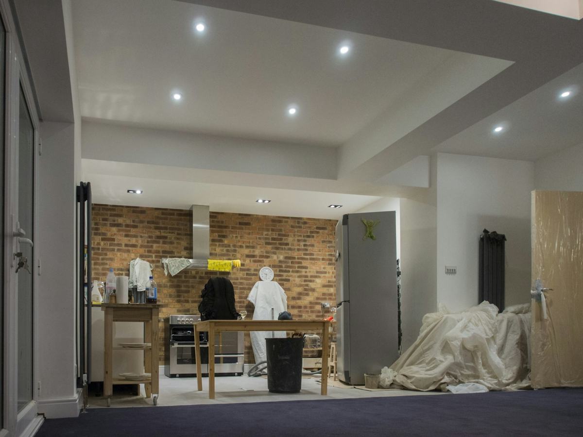 Room under renovation with furniture covered in dust sheets and workers in protective coveralls.